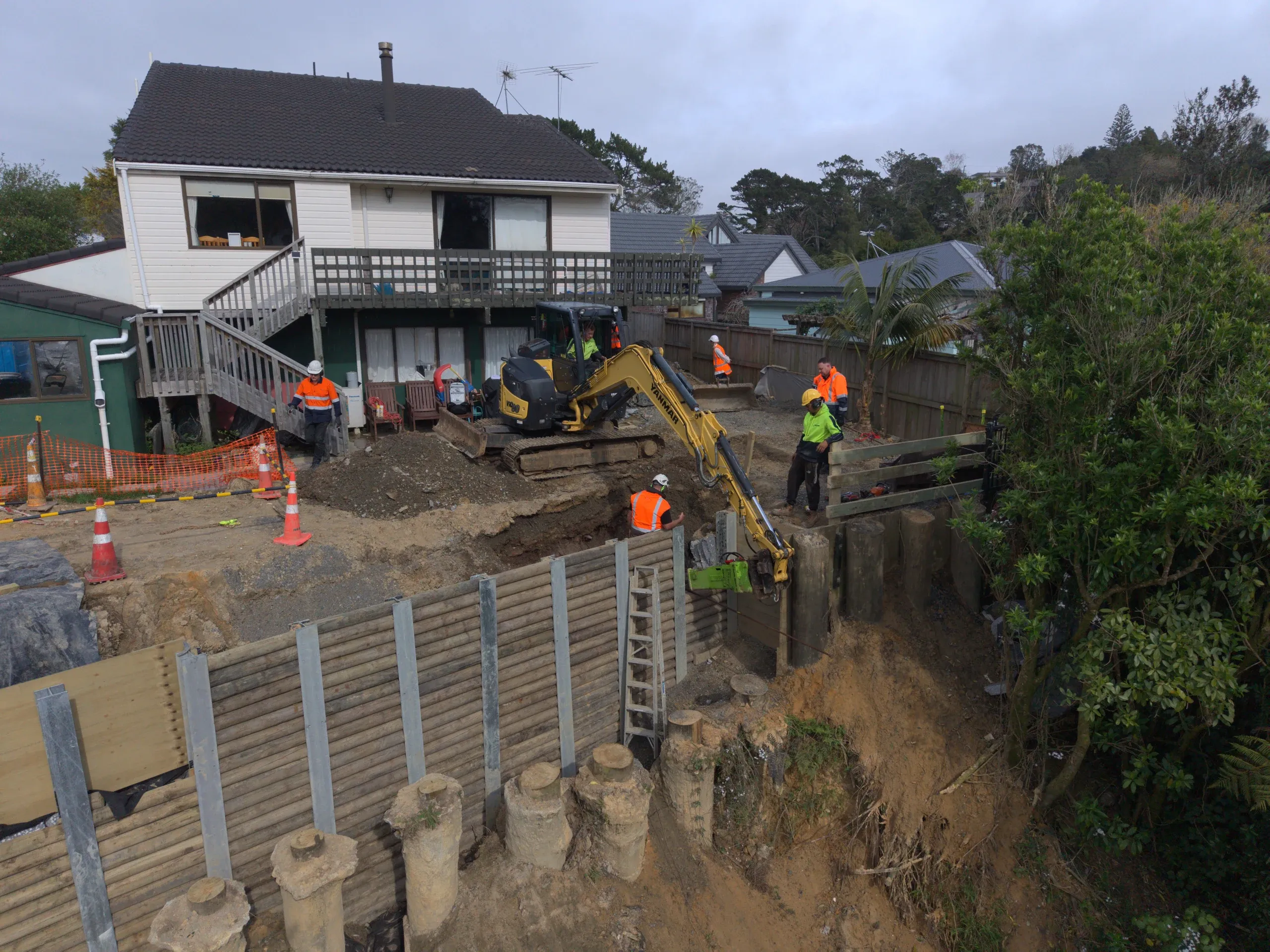Aerial view of the excavator working the pile line, with exposed pile heads and the retaining wall taking shape below the house.