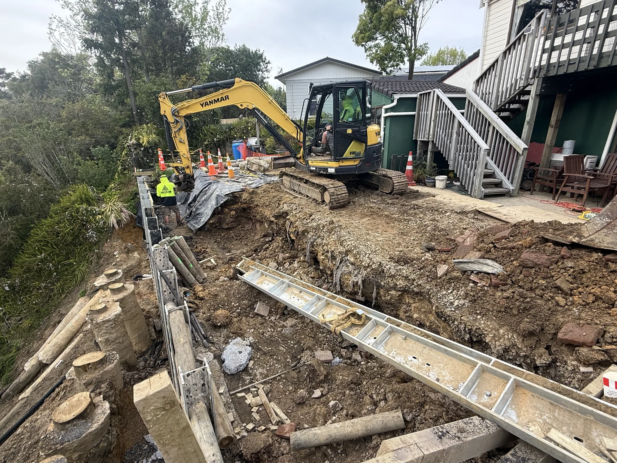 Excavator on the cut platform beside the part-built retaining wall, with a steel channel laid out ready to install.