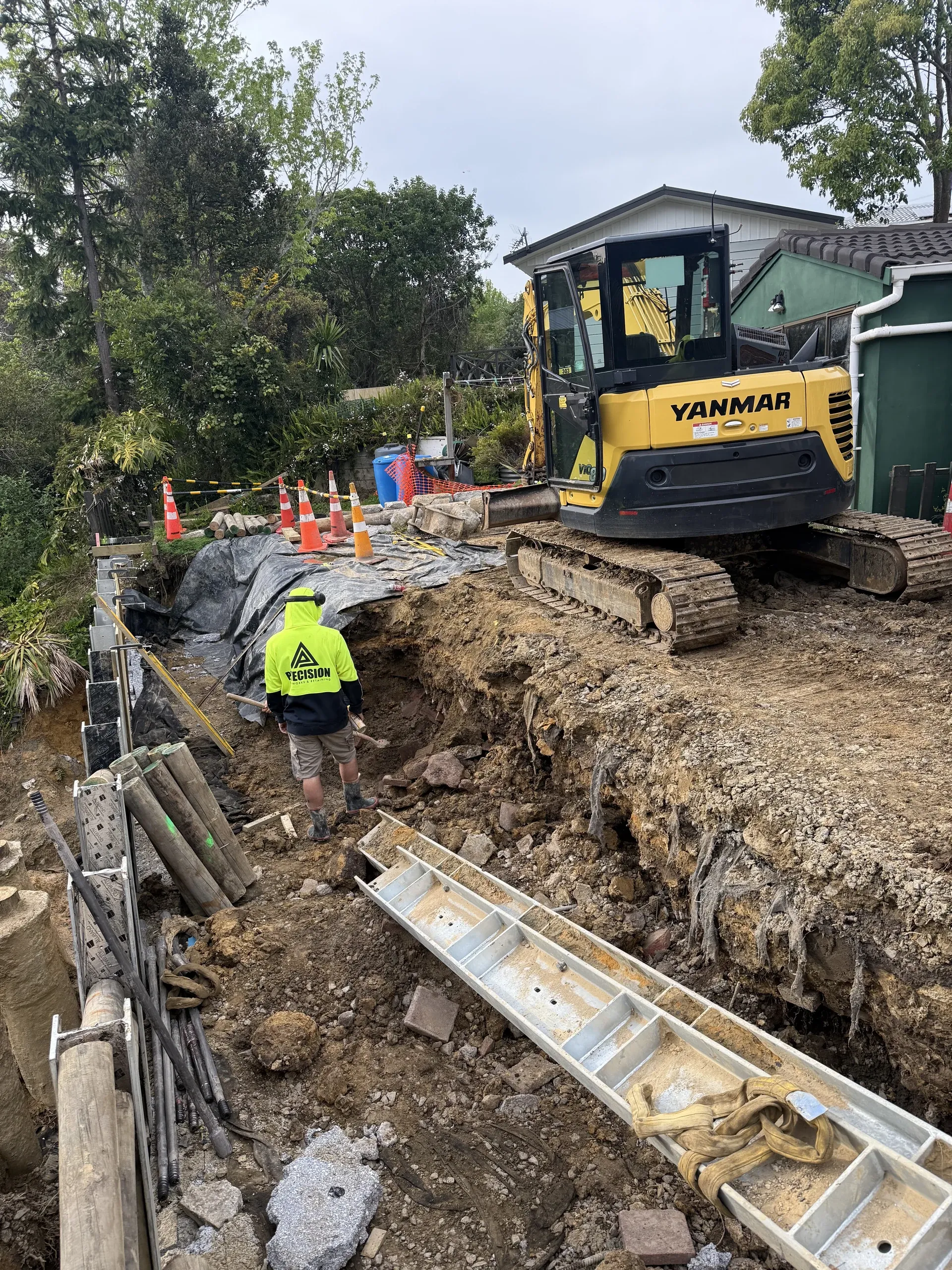 Crew member working the wall line beside the excavator, with steel channel and pile stock stacked on the platform.