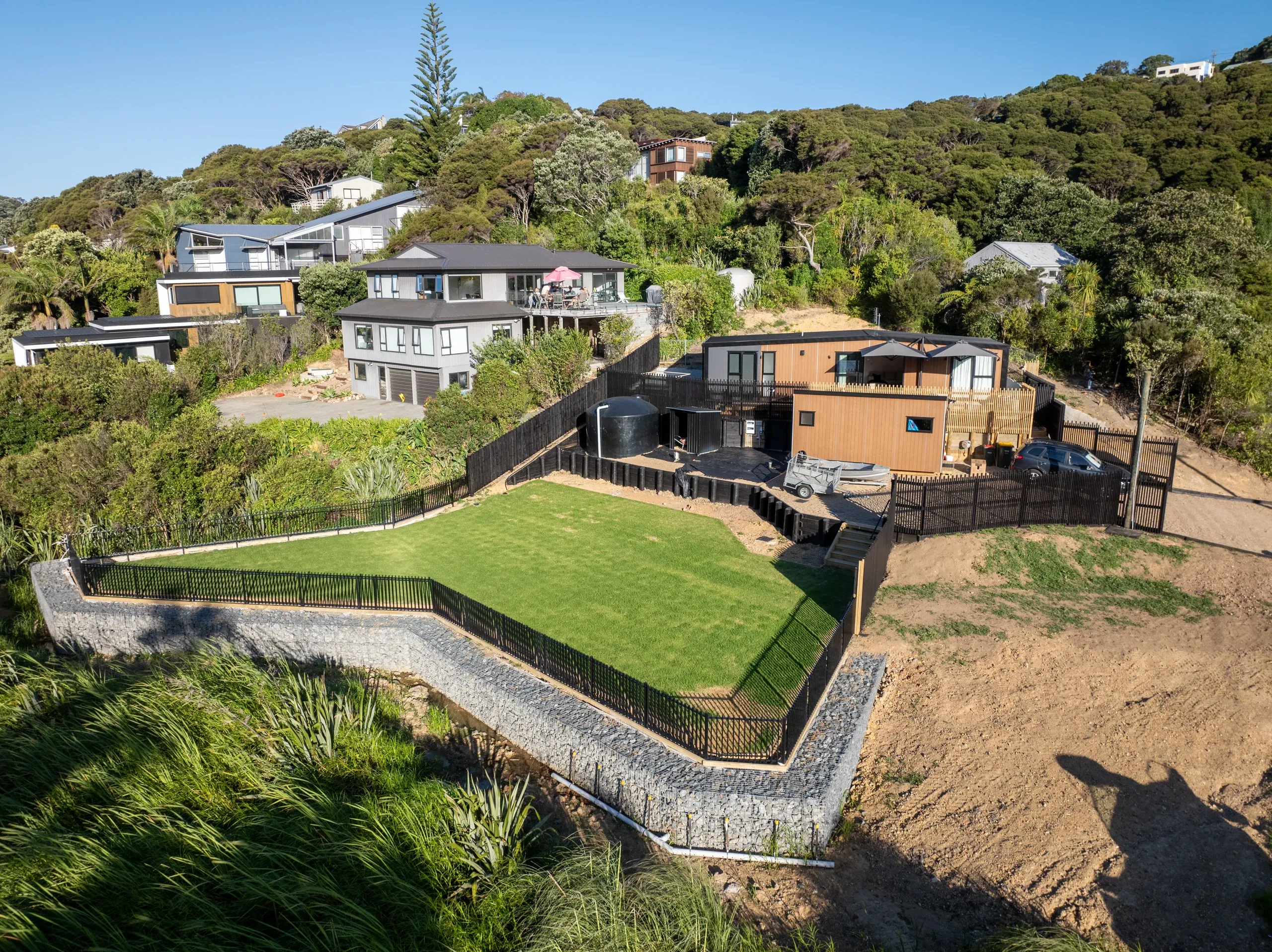 Aerial view of the completed Waiheke terrace: a level lawn held by gabion retaining, with the house and coastal bush beyond.