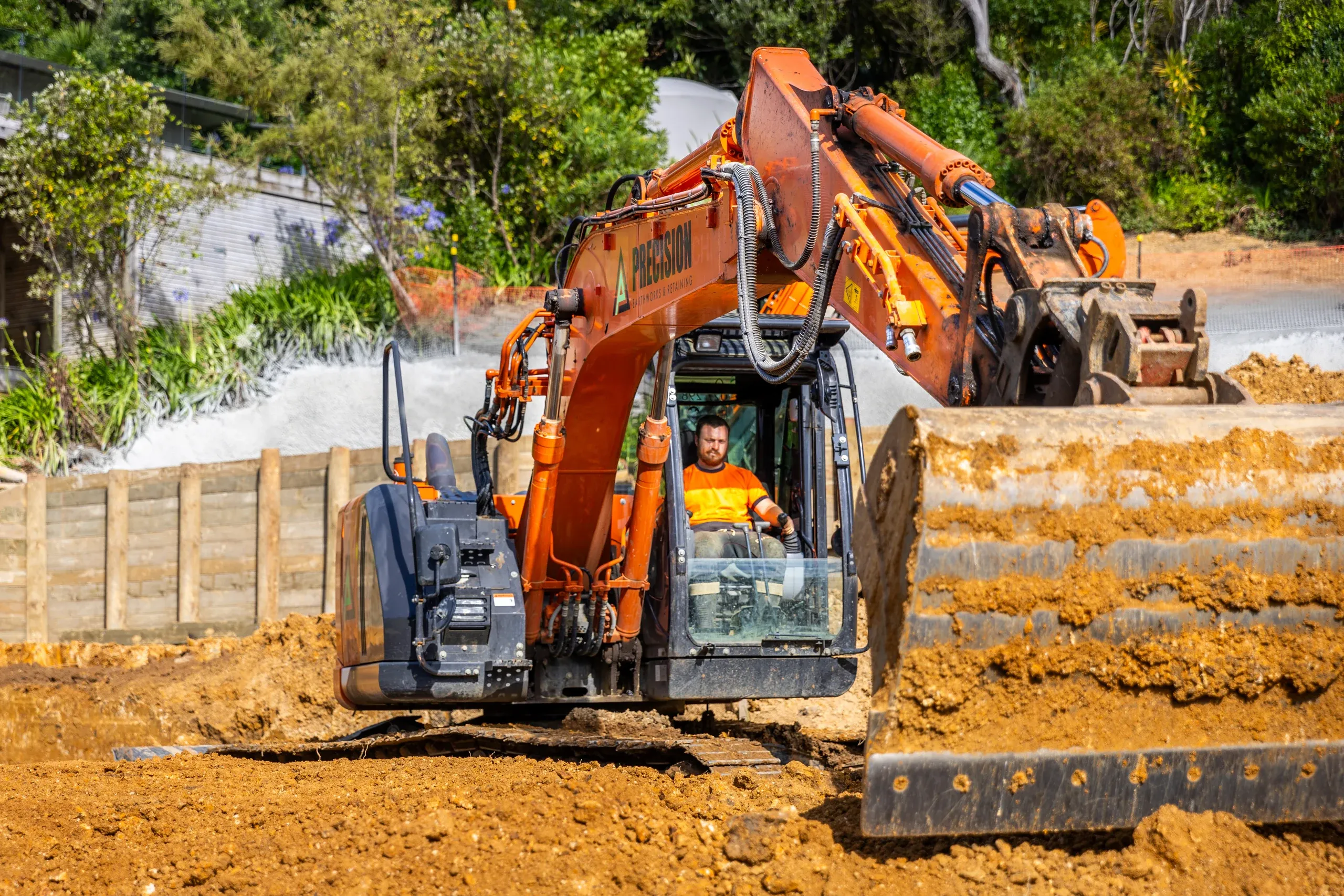 The excavator tracking along the completed timber pole retaining run, terraced ground stepping away behind.