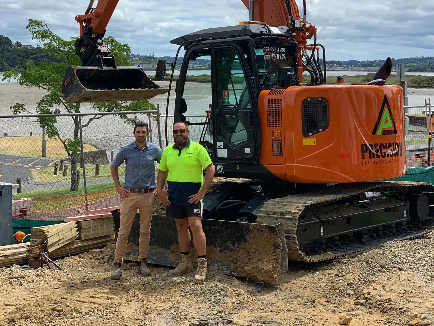 Two Precision Earthworks team members standing in front of a branded Hitachi excavator on a coastal Auckland site