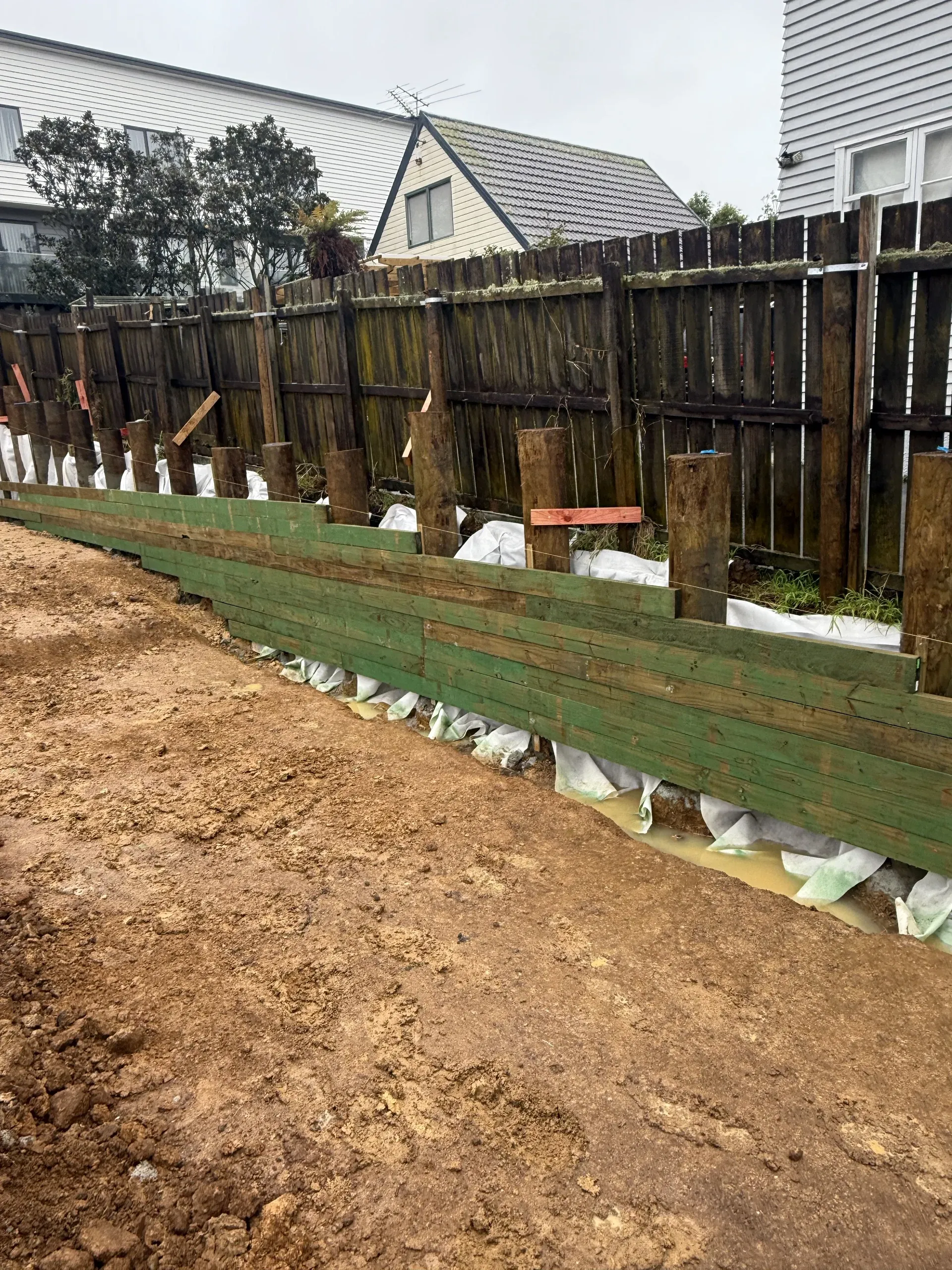 Timber retaining wall going up against the boundary fence, with geotextile-wrapped drainage laid in behind the rails.